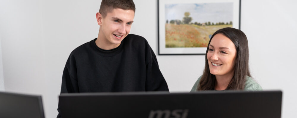 Zwei junge Mitarbeitende arbeiten gemeinsam an einem Computer und tauschen sich lächelnd aus. Das Bild vermittelt eine moderne Arbeitsumgebung, starke Teamdynamik und die Förderung von Nachwuchskräften in der Steuerkanzlei von Karin Klehm.